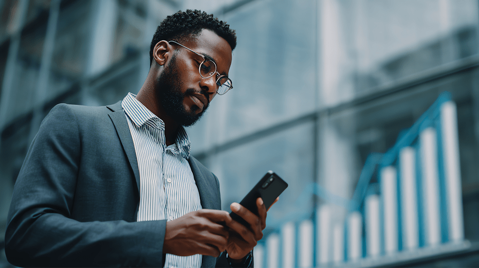 Black man in glasses wearing business clothes looks at a phone in front of a building with a bar graphing showing upward growth behind him.